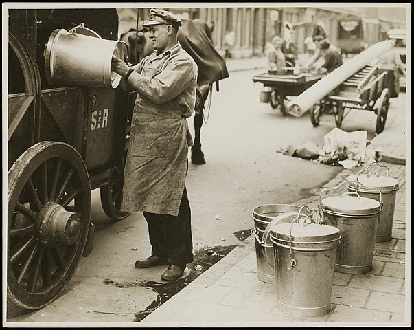 Foto van een vuilnisman in Amsterdam die een vuilnisemmer in een vuilniswagen leegt; circa 1930.